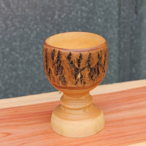 May include: A wooden goblet with a natural wood finish and a bark-like design around the top. The goblet has a rounded base.