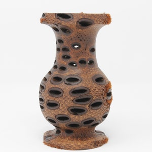May include: A small, brown, wooden vase with a unique design. The vase is made from a natural material with a pattern of holes throughout. It has a rounded body and a flared top.