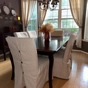 May include: A dining room set with a dark wood table and four chairs covered in white slipcovers. A chandelier hangs above the table, and the room has large windows with cream-colored curtains. A vase of flowers sits on the table.
