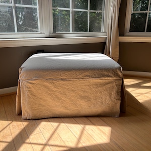 May include: A square ottoman with a light beige slipcover. The ottoman is in a room with hardwood floors and large windows. The sunlight casts shadows on the floor and the ottoman.