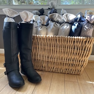 May include: Two black leather boots with buckles, sitting in front of a wicker basket filled with fabric pouches. The pouches are in various shades of gray and white.