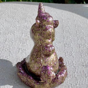 Pu&ograve; includere: Una statuetta di unicorno dorato e viola glitterato. L'unicorno &egrave; seduto con la testa girata, con un corno a spirale e una criniera di glitter viola. La statuetta ha una superficie strutturata.