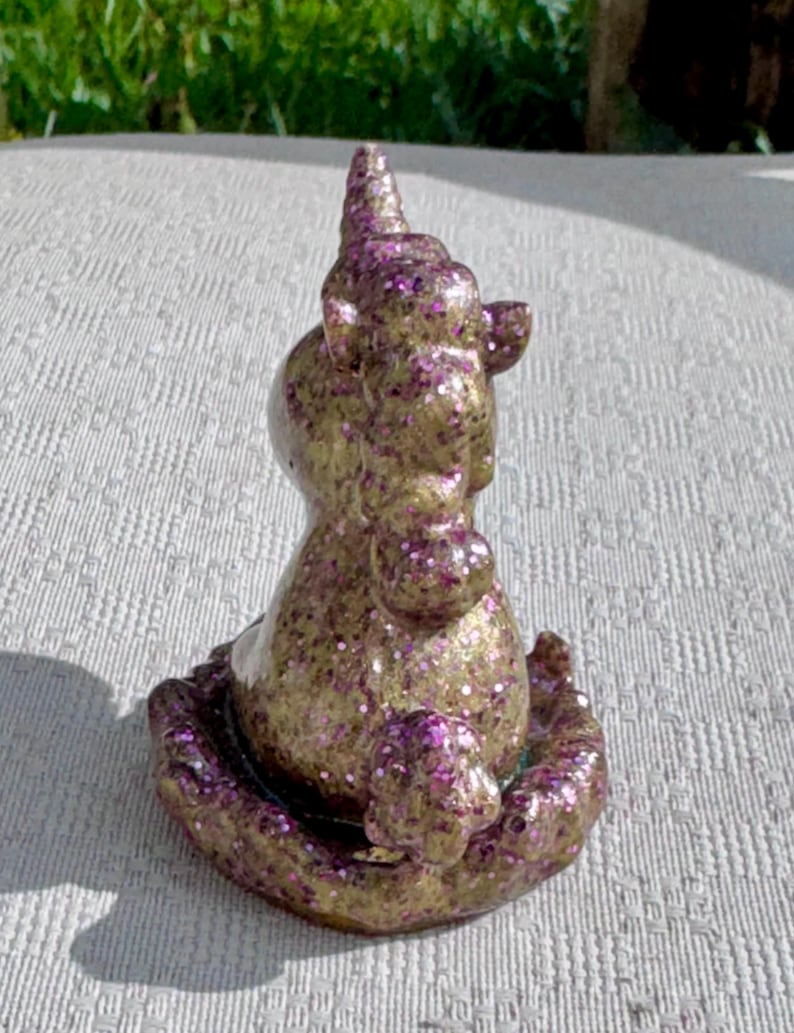 Pu&ograve; includere: Una statuetta di unicorno dorato e viola glitterato. L'unicorno &egrave; seduto con la testa girata, con un corno a spirale e una criniera di glitter viola. La statuetta ha una superficie strutturata.