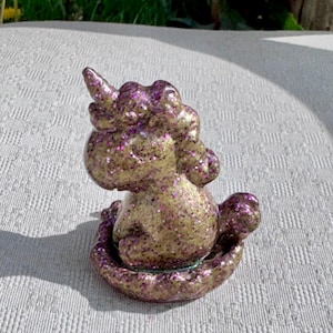 Pu&ograve; includere: Una statuina di unicorno dorato e viola glitterato. L'unicorno &egrave; seduto, con un corno prominente e una criniera. La statuina &egrave; ricoperta di glitter viola, che le conferiscono un aspetto scintillante. Anche la base &egrave; viola e glitterata.