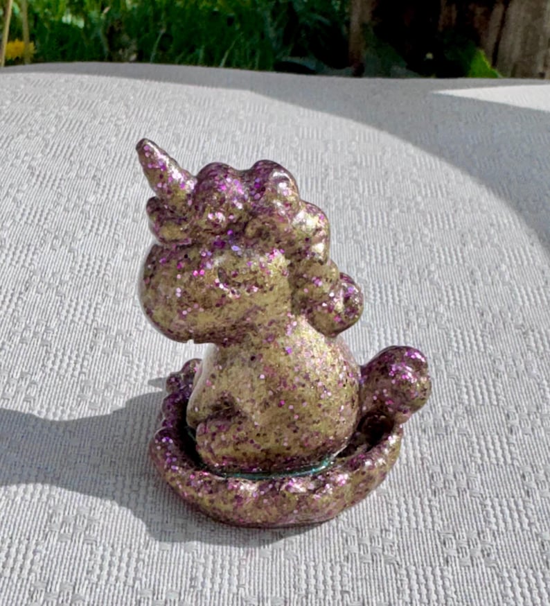 Pu&ograve; includere: Una statuina di unicorno dorato e viola glitterato. L'unicorno &egrave; seduto, con un corno prominente e una criniera. La statuina &egrave; ricoperta di glitter viola, che le conferiscono un aspetto scintillante. Anche la base &egrave; viola e glitterata.
