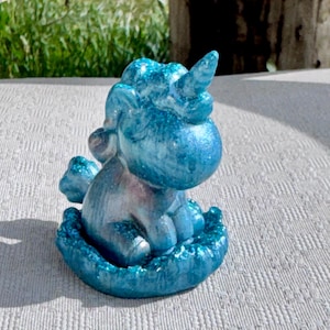 Pu&ograve; includere: Piccola statuina di unicorno blu glitterato. La creatura mitica &egrave; seduta, con il ventre argentato e un singolo corno. La statuina poggia su una base strutturata, con una finitura scintillante. Lo sfondo &egrave; sfocato, con erba verde e un palo di legno.