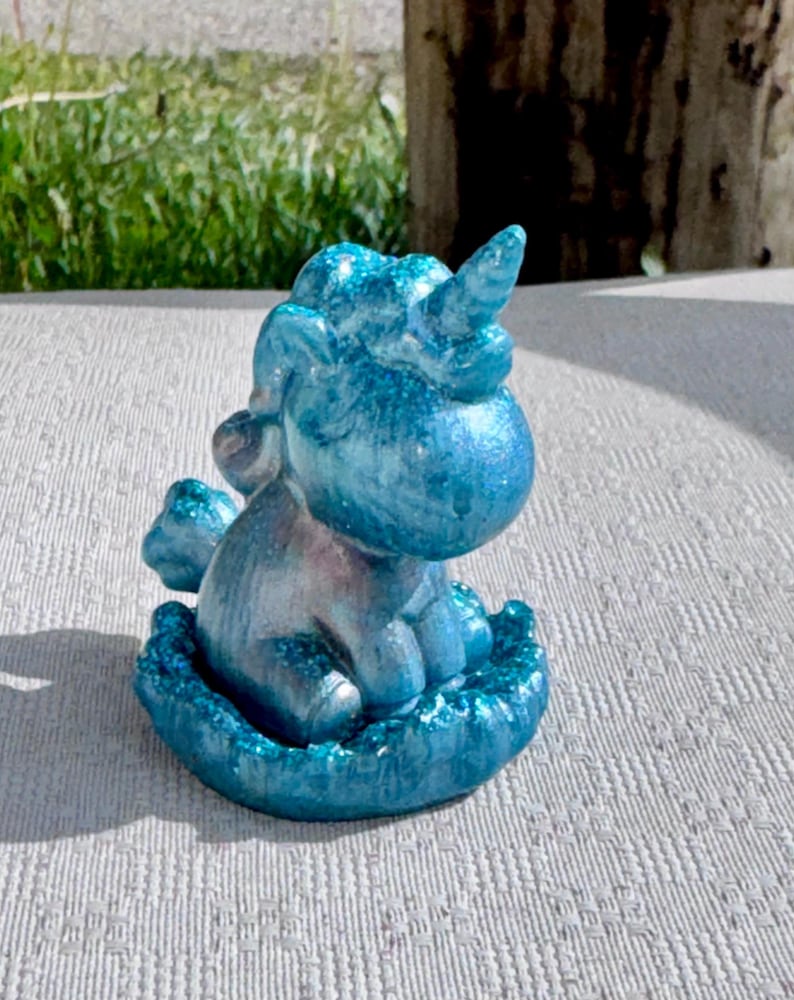 Pu&ograve; includere: Piccola statuina di unicorno blu glitterato. La creatura mitica &egrave; seduta, con il ventre argentato e un singolo corno. La statuina poggia su una base strutturata, con una finitura scintillante. Lo sfondo &egrave; sfocato, con erba verde e un palo di legno.