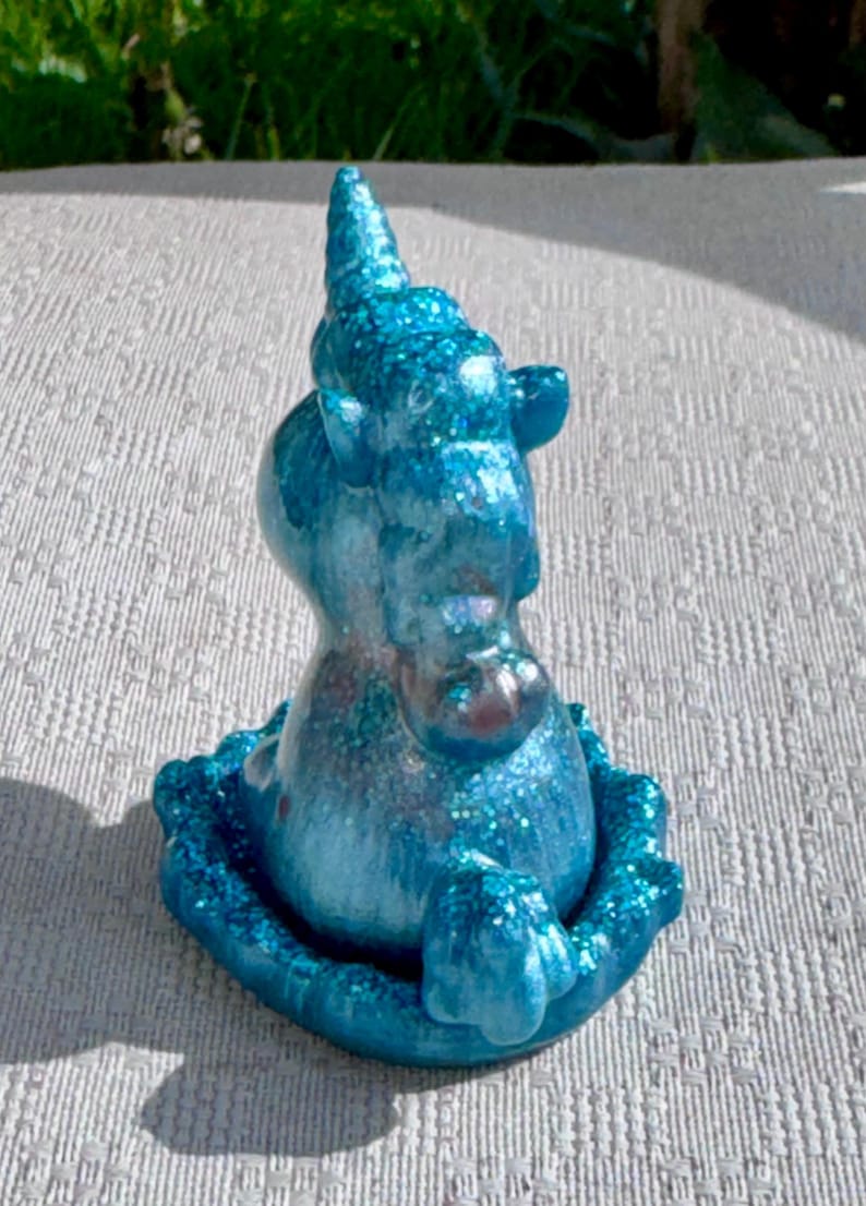 Pu&ograve; includere: Piccola statuetta di unicorno blu glitterato. L'unicorno ha un corno a spirale ed &egrave; seduto su una base decorativa. La statuetta &egrave; di una vibrante tonalit&agrave; di blu con accenti glitterati scintillanti, creando un'estetica stravagante e magica.