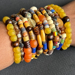 May include: A collection of beaded bracelets in various colors, including yellow, brown, orange, and blue. The bracelets feature a mix of wooden, glass, and metal beads, creating a layered and textured look. The bracelets are stacked on top of each other.