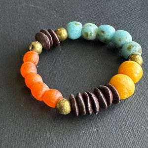 May include: A beaded bracelet featuring a mix of orange, yellow, turquoise, and brown beads. The bracelet includes round and disc-shaped beads in various sizes and textures, strung together with gold-colored spacers. The bracelet is displayed on a dark surface.
