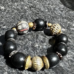 May include: A beaded bracelet featuring a mix of black, gold, and patterned beads. The bracelet includes large, round black beads interspersed with smaller gold-toned and patterned beads. The design is a mix of textures and colors.