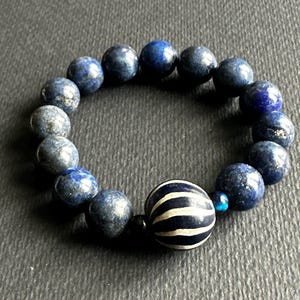 May include: A beaded bracelet featuring dark blue lapis lazuli beads and a single, larger striped bead in navy and white. The bracelet is strung on an elastic band, creating a circular shape.