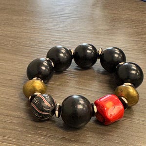 May include: A beaded bracelet featuring large, round black beads, interspersed with gold-colored and patterned beads. A single red, square bead adds a pop of color. The bracelet is displayed on a wooden surface.