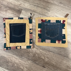 May include: Two square quilted pot holders. Each features a denim pocket as a central design element. The pockets are surrounded by a tan border and a patterned fabric border with green, red, and gold accents. Brown loops are attached for hanging.