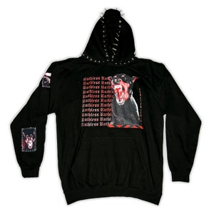 Puede incluir: Sudadera con capucha negra con un estampado gráfico de un perro gruñendo y el texto repetido "Ruthless Ruth" en rojo. La sudadera tiene tachuelas plateadas alrededor de la capucha y en las mangas.