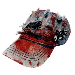 May include: A red and blue denim baseball cap with a distressed look. The cap has a patch with the word "RIP" on it and is decorated with silver spikes. The cap has a red and white splatter pattern.