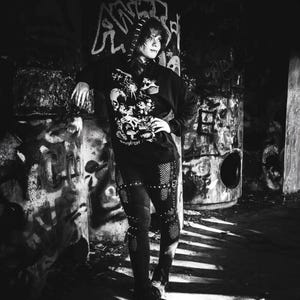 May include: A black hooded sweatshirt featuring a white graphic print of a skull, spider, and mushrooms with the text "Necrycrypt" on the front. The sweatshirt is paired with black pants that have integrated fishnet panels and straps.