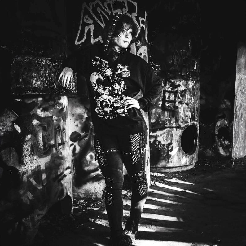 May include: A black hooded sweatshirt featuring a white graphic print of a skull, spider, and mushrooms with the text "Necrycrypt" on the front. The sweatshirt is paired with black pants that have integrated fishnet panels and straps.