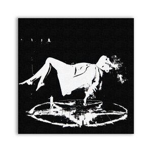 Può includere: Un'immagine in bianco e nero di una persona che levita sopra un pentagramma. La persona indossa una lunga tunica fluida e ha le braccia distese. Il pentagramma è disegnato sul pavimento e ci sono candele intorno.