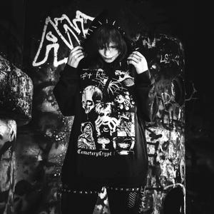 May include: A black hooded sweatshirt featuring a white graphic print of a skull, a hooded figure, a mushroom cloud, and a snarling creature. The hood is adorned with metal spikes. The text CemeteryCrypt is printed in white on the front.