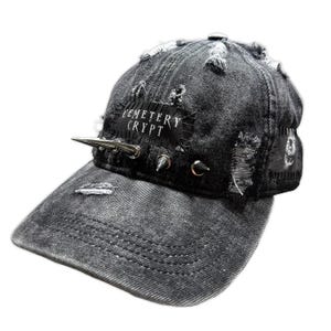 May include: Distressed black denim baseball cap with silver spikes and the text "CEMETERY CRYPT" on a black patch. The cap has a worn, ripped design, giving it a punk rock aesthetic. The brim is curved and the cap is a standard size.