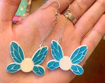 Pode incluir: Um par de brincos prateados com um design de borboleta. As asas da borboleta são turquesa com um padrão detalhado e um centro circular branco. Os brincos penduram de uma corrente e um gancho.