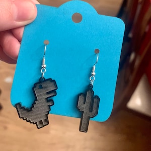 May include: A pair of black pixelated earrings. One earring is a dinosaur, the other is a cactus. The earrings hang from silver-coloured fish hook ear wires. The earrings are displayed on a light blue card.