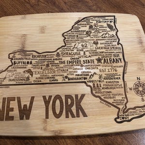 May include: A wooden cutting board with a laser-engraved map of New York State. The map includes major cities, landmarks, and geographical features. The text "NEW YORK" is prominently displayed below the map.