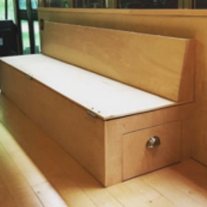 Könnte beinhalten: Eine helle Holzbank mit einem Klappdeckel, der sich öffnet und Stauraum freigibt. Die Bank hat eine kleine Schublade mit einem silbernen Knopf an der Vorderseite.