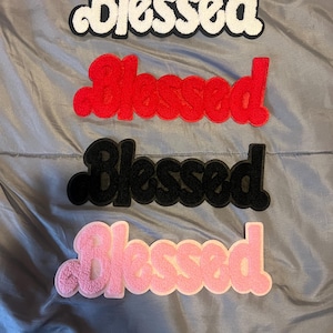 May include: Four "Blessed" patches in different colors: white, red, black, and pink. The patches are made of fuzzy fabric and have a slightly raised, 3D effect.