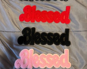 May include: Four "Blessed" patches in different colors: white, red, black, and pink. The patches are made of fuzzy fabric and have a slightly raised, 3D effect.
