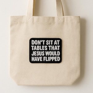 May include: A beige tote bag with a black rectangular patch. The patch features white text that reads: "DON'T SIT AT TABLES THAT JESUS WOULD HAVE FLIPPED."
