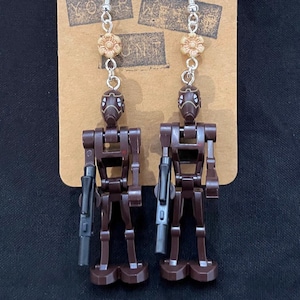 May include: A pair of handmade earrings featuring dark brown robot figures with silver hooks. Each earring has a small, tan flower bead and a silver chain. The earrings are displayed on a brown card with the text "YOUR WEAKNESS".