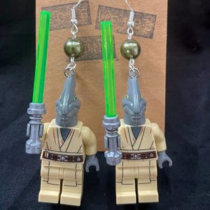 Peut inclure: Boucles d'oreilles faites à la main avec des figurines Lego. Elles présentent des têtes grises, des corps beiges et des sabres laser verts. Les boucles d'oreilles ont des crochets argentés et des perles vert olive. Présentées sur une carte marron.