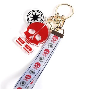 Pode incluir: Um chaveiro de caveira vermelha com um amuleto de símbolo imperial branco e preto e uma fita branca com desenhos repetidos de caveira vermelha e símbolo imperial.