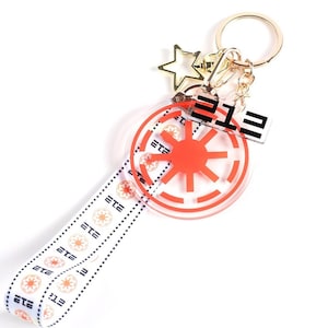 May include: A gold-colored keychain with a clear, round charm featuring a red and white design. The keychain includes a strap with a repeating pattern and the text "ETE". A small star charm hangs from the chain.