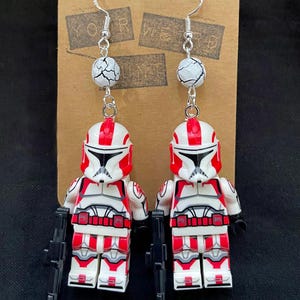 May include: A pair of dangle earrings featuring miniature red and white toy soldier figures with black weapons. The earrings have silver hooks and are accented with white and gray marbled beads. The earrings are displayed on a brown cardboard backing.