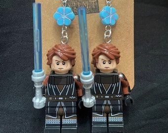 May include: A pair of handmade earrings featuring Lego figures of a character with a lightsaber. The earrings have blue flower charms and the text "YOUR WEIRD UNIT" on the backing card. The figures are brown, black, and gray.