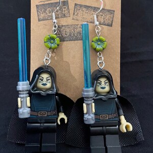 May include: A pair of earrings featuring a Lego minifigure of a female character from Star Wars holding a blue lightsaber. The minifigure is wearing a black robe and has green skin. The earrings are silver and have a green flower charm.