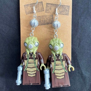 May include: A pair of dangle earrings featuring a green and brown Lego minifigure of a character from Star Wars. The minifigure is wearing a brown robe and has a green head with a breathing apparatus. The earrings are silver and have a round bead at the top.