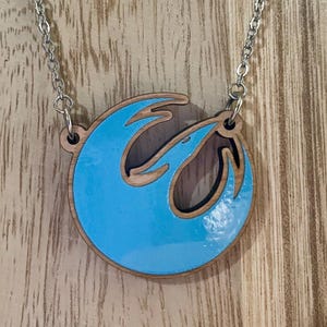Könnte beinhalten: Eine silberne Kettenkette mit einem blauen und braunen Holzanhänger mit dem Symbol der Rebellenallianz aus Star Wars.
