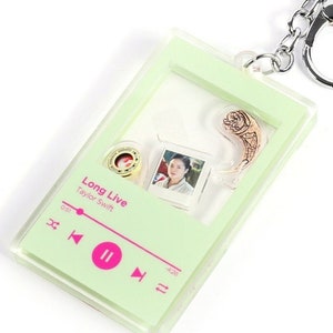 May include: A green and clear acrylic keychain with a music player design. The keychain features a picture of Taylor Swift, the song title "Long Live", and the song length "0:31 - 4:26".