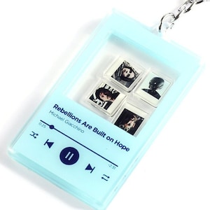 May include: A light blue keychain shaped like a music player with a clear window showing four small photos. The text on the keychain reads "Rebellions Are Built on Hope" and "Michael Giacchino".