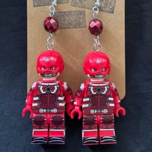 May include: A pair of novelty earrings with red Lego minifigure charms, silver-tone hooks, and red faceted beads. The minifigures have red helmets and detailed red and white outfits. The earrings are displayed on a brown card.