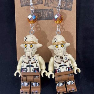 May include: A pair of dangle earrings featuring Lego minifigures of a Star Wars character, a Geonosian, with amber colored glass beads. The earrings are on a brown card with the text "Your Weird Aunt".