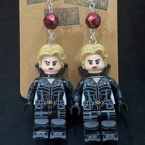 May include: A pair of dangle earrings featuring Black Widow Lego figures. Each earring has a silver hook, a faceted red bead, and a Black Widow Lego figure with a blonde hairstyle and black outfit. The earrings are displayed on a brown card.