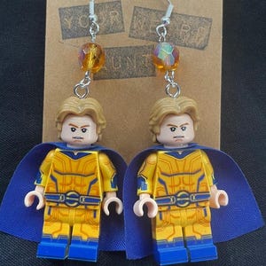 May include: A pair of handmade earrings featuring Lego minifigures with yellow and blue superhero costumes. The earrings have silver hooks and are accented with orange crystal beads. The minifigures have blonde hair and blue capes.