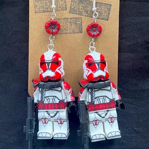 Może przedstawiać: Para ręcznie robionych kolczyków z miniaturowymi białymi i czerwonymi figurkami z czarną bronią. Kolczyki mają srebrne haczyki i są ozdobione czerwonymi koralikami w kształcie kwiatów. Figurki są przymocowane do brązowej karty.