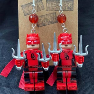 May include: Handmade earrings with Lego-style superhero figures in red and black, each holding a silver trident. The earrings have red bead accents and silver hooks. The figures have a red cape and the letter "D" on the chest. The background has the words "Your Weird Aunt".