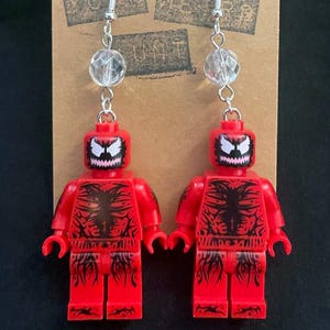 May include: A pair of red Lego-style earrings featuring a character design with a white face and black detailing. Each earring has a clear, faceted bead and silver-tone ear wire. The earrings are displayed on a brown card with text.